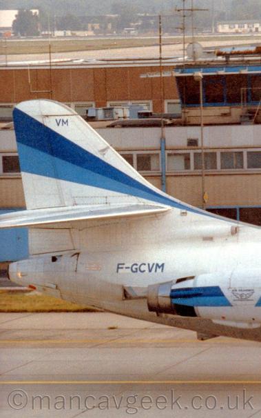 Close up of the white rear fuselage and tail of a twin engined jet airliner facing to the right with the engines mounted on the sides of the rear fuselage.
The tail has a two-tone blue running from the lower front edge to the top rear, getting wider as it goes.
A similar stripe runs along the engine pods, with a white section in the middle with a winged logo with "Air Provence" title above it.
The registration "F-GCVM" is on the upper rear fuselage, just aft of the engines, in blue.
A wide building in shades of brown stretches across the background, with trees and low buildings in the distance.