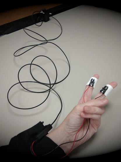 A hand showing a "V" sign. Galvanic skin response electrodes are placed on the hand's index and middle fingers. They are wired and the cable leads to a wooden wall.
