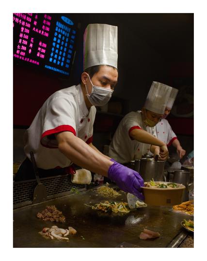 This image shows a scene of chefs working in a restaurant or street food setting. The main focus is on a chef wearing a tall white chef hat, a white uniform with red trim, and a face mask. He is also wearing a purple glove on one hand as he prepares food on a large, flat griddle. The griddle has various ingredients on it, including what appears to be meat, seafood, and vegetables. 

The chef is placing cooked food into a bowl, likely for serving. In the background, another chef wearing a similar uniform and hat is also busy preparing food. Behind them, there is a menu board with Chinese characters written in bright neon colors, indicating the dishes and prices available. The overall lighting is dim, creating a cozy atmosphere, and the focus is on the active work being done by the chefs.