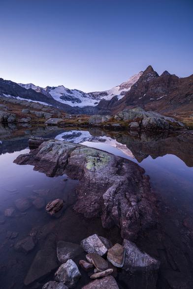 Photo d'un paysage de montagne.
De hautes montagnes se reflètent dans un petit lac. Certains pics sont saupoudrés de neige, on constate aussi la présence d'un glacier à leur pied.
Une partie du petit lac est encombrée par un gros rocher et quelques galets, masquant une partie du reflet.
Le ciel est bleu mais le jour se lève à peine, seul un sommet est faiblement éclairé. Une partie du paysage ainsi que les rochers au premier plan ont une teinte violacée, due à la lumière du lever du jour.
-
Photo of a mountain landscape.
High mountains are reflected in a small lake. Some peaks are dusted with snow, and a glacier can be seen at their foot.
Part of the small lake is blocked by a large boulder and a few pebbles, masking part of the reflection.
The sky is blue, but daylight is just breaking, with only one summit dimly lit. Part of the landscape and the rocks in the foreground have a purplish hue, due to the early morning light.
