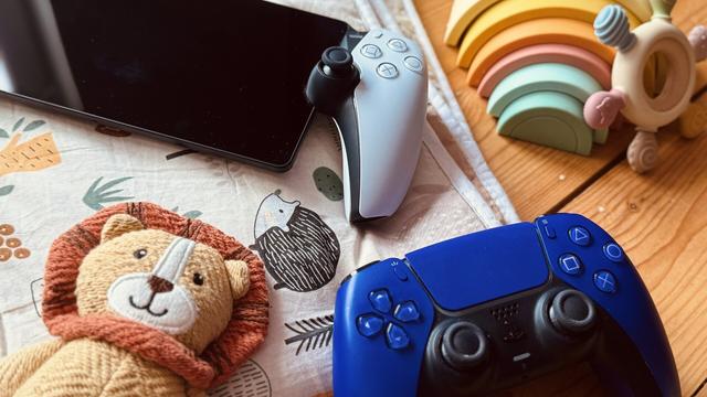 The picture shows a PlayStation controller and the PlayStation Remote player on a wooden background, next to a stuffed lion, a rainbow stick figure and a chew toy. The combination of gaming accessories and baby toys symbolizes the interplay between gaming and parenthood.