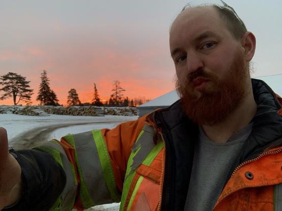 A hefty white cow male offers out a hug. He has a bright red beard and a reflective jacket. Behind him the sun is slowly rising