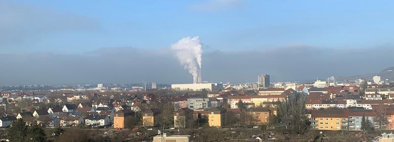 A panoramic city view features residential buildings with varied architecture in the foreground, while a large industrial facility with smokestacks emitting white smoke is visible in the background against a clear, blue sky.