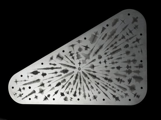 One side of the silver-gray, rounded, triangular 'Water Words' plate with black engraved waveforms arranged in an radial  fashion.