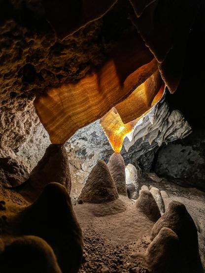 Inside a cave, thin ribbons of rock are illuminated from behind revealing delicate bands varying in color from brown to amber.  The lower tips of these ribbons merge with gumdrop-shaped mounds of rock rising from the cave floor.