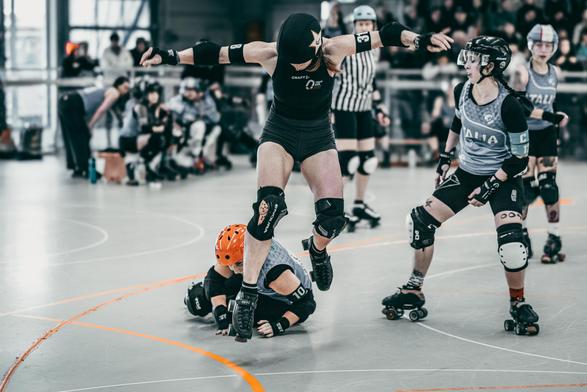 A color photo of a Roller Derby Jammer that is jumping over another player, arms spread wide