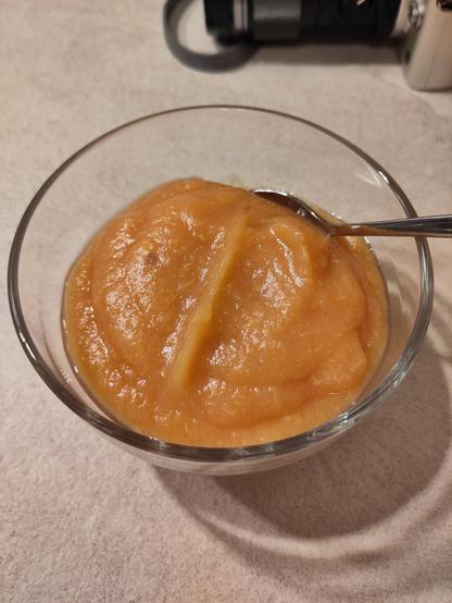 Das fertige Quittenmus in einem kleinen Glasschälchen. Am Rand steckt ein Teelöffel und im Hintergrund ist eine Kamera zu sehen mit der gerade Fotos vom Mus für den Foodblog gemacht wurden.