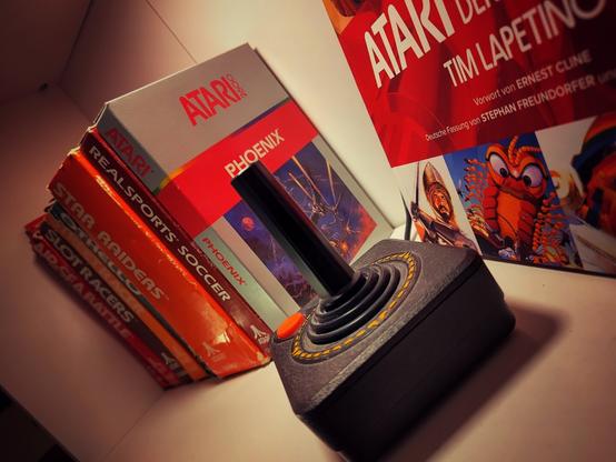 An Atari CX-40 joystick in a shelf. Behind it some game boxes of Atari 2600 cartridges. On the right side the book "The Art of Atari" from Tim Lapetino is lurking into the scene.