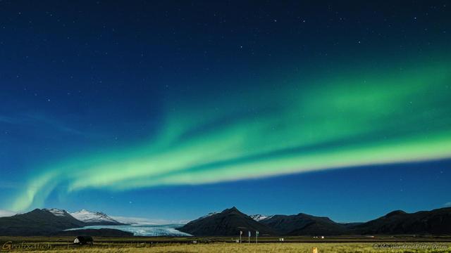 A colour photo of a night scene with a dark sky full of stars. Six of the seven stars of The Plough or Big Dipper, properly known as Ursa Major, are visible in the top right. The foreground is flat grassland with some flagpoles in the near distance. Beyond them is a mountain range and, dropping between them, a glacier folding down to ground level. Above and seeming to come from the glacier  itself are stripes of green light, some in quite straight lines, others swirling up to another level. The base of some of the ribbons is yellowish, and the colours blend and fade in patterns.