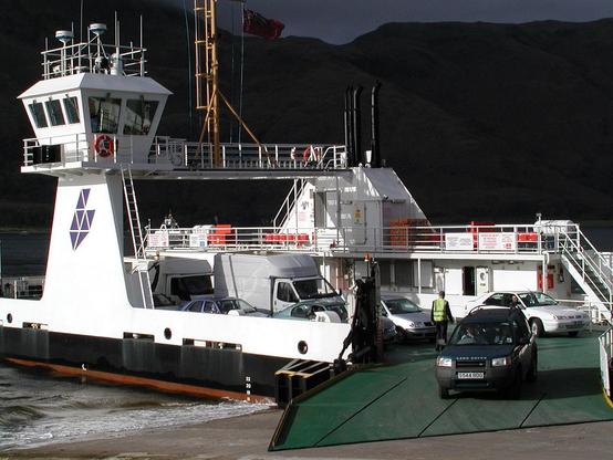 The Corran Ferry. The image shows the ferry in bright sunshine with vehicles driving off, towards the camera. The background is very dark, creating a striking contrast. The ferry has a white superstructure above a black hull and the vehicle deck is green.
