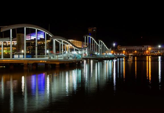 Una larga exposición nocturna del puente con los reflejos de las luces en el agua del mar del centro comercial Maremagnum, en Barcelona, España.

A long exposure night photo of the bridge with the reflections of the lights in the seawater of the Maremagnum shopping center, in Barcelona, Spain.