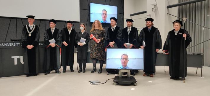 Me and my PhD-committee lined up looking towards a photographer:

From left to right: Barry Koren as chair, Tibor Jager, Peter Schwabe, Tanja Lange as 2nd promotor, me, Andreas Hülsing as 1st promotor, Berry Schoenmakers, Benjamin Dowling and an assistant of TU Eindhoven. In front (and behind) of us a screen with Kenny Patterson who joined remotely.

I wear a midi-length black dress with chaotic yellow lines all over it, holding a round box that contains my certificate. Everyone else who is present in person wears a black gown and a black hat, except for Tanja who wears a black suit, because she thinks the gowns look ridiculous and a face-mask.