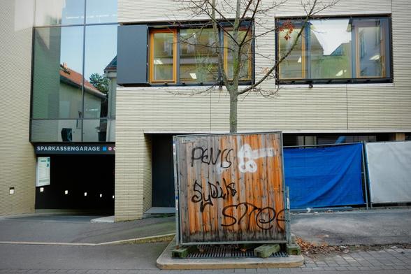 Ein Holzzaun mit Graffiti vor einem modernen Gebäude. Das Graffiti zeigt verschiedene Schriftzüge und ein weißes Symbol. Im Hintergrund ist ein Gebäude mit großen Fenstern und einem Schild mit der Aufschrift "SPARKASSENGARAGE" zu sehen.
