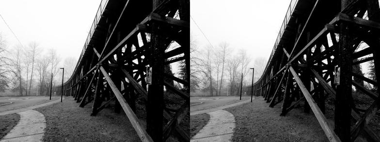 Black and white, stereoscopic, side-by-side image. A view from the bottom of a former train trestle, currently supporting a pedestrian bridge. The wooden structure is mostly on the right side of the image, receding toward the center of the image, into the fog.