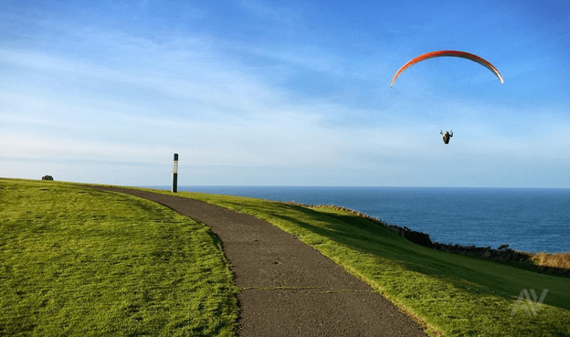 En la foto puede verse un trozo de camino en el monte de San Pedro, rodeado de hierba. Puede verse también el cielo azul con unas pocas nubes, una persona volando en parapente, y unas trozo de mar.