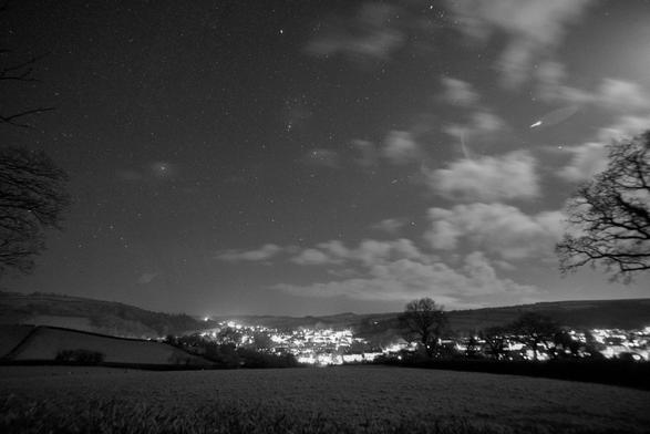 Monochrome night-scape. Foreground is grass fields, sloping down and away from the viewpoint into a valley. The valley, in the centre contains dazzling and numerous town lights. In the far distance, behind the town, gentle hills. The far left and right of the view have bare tree branches which frame the sky. The night sky is split roughly in half with motion blurred, bright white, moonlit clouds on the right, and many stars on the left. Orion is in the centre with Canis Major on the left. Sirius appears somewhat subdued due to a small patch of cloud. There is a bright lens flare on the cloudy side of the sky from the moon, which is out of frame.