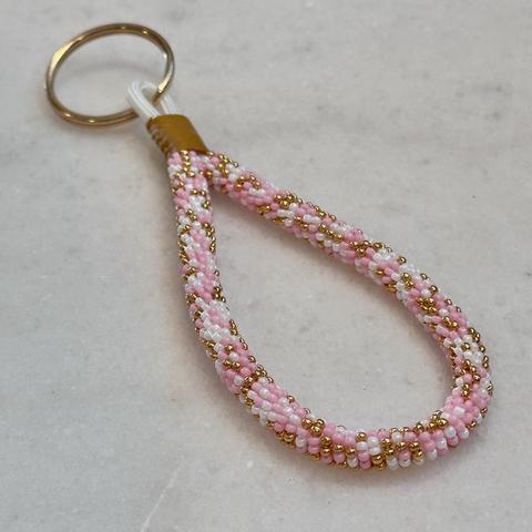 A bead-wrapped mini keychain in blush pink, gold, and pearl white, with a gold split ring, on a marble background