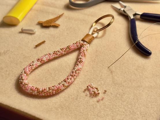 A bead-wrapped keychain in blush pink, gold, and pearl white, on a bead mat

You can see materials and tools around the bead mat such as pliers, needle, scraps of leather, and seed beads.