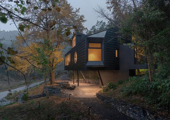 A modern, elevated dark-colored house with large windows is set amidst a forested area. The house is lit inside, and a small path leads up to it.