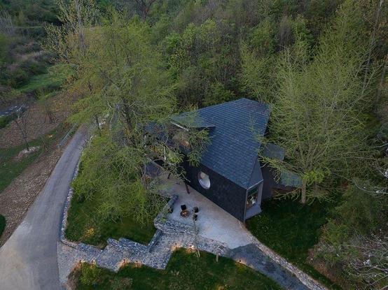 Aerial view of a modern, dark-colored house with a sloped roof, situated in a wooded area. A stone pathway and small patio are visible amidst surrounding trees and greenery.