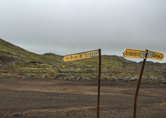 In the foreground, two weather-beaten signposts planted in the ground. One stands straight, pointing left and reading "Hellissandur 20 [F575]". The second is bent midway up, with two arrows: poining left and back reads "Arnarstapi 13 [570]"; pointing rightward reads "[570] Olafsvik 10". Behind them, a rocky mountainside, with small mosses and lichen covering the rocks in the distance. The sky is overcast.