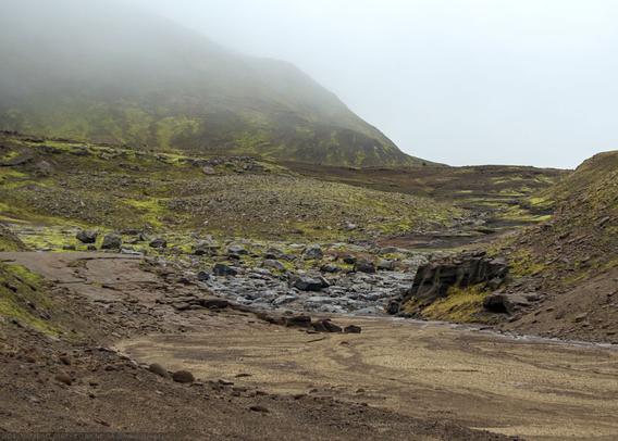 In the foreground, a dry, sandy glacial riverbed. Further on, larger loose rocks, and beyond a mountain covered in thin moss and lichen vanishes into the low clouds.