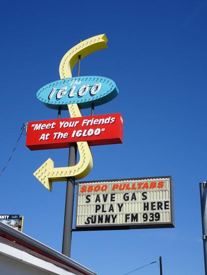 A retro midcentury sign against a blue sky with a large yellow line swooping downward in an s-shape with an arrow at its end pointing leftward. Toward the top of the line is a turquoise blue oval that says "the Igloo." Further down is a red rectangle that says "Meet your friends at the Igloo." Beneath the arrow is a another sign that says "$500 pulltabs. Save gas. Play here. Sunny FM 939."