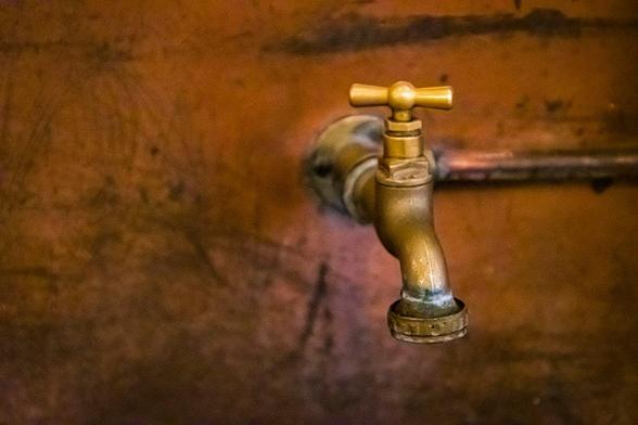 The image shows a brass faucet attached to a wall. The background appears to be made of a brown material, possibly wood or metal. The faucet has a traditional design with a cross-shaped handle.
