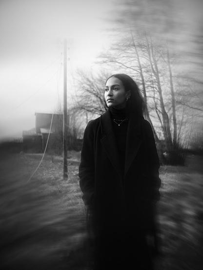 Portrait of a young woman in a long black coat standing somewere in a rural area and looking to the side. The edges of the image are very blurred.