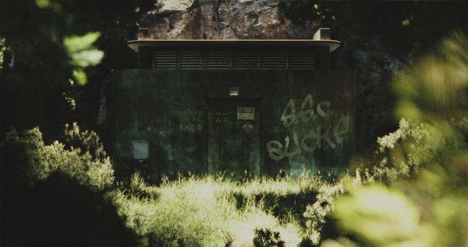 A picture of Stelpolvo Lab super collider emergency exit 6 on the densely wooded hill of the crater lake near Okulago. The small building is made of concrete with copper cladding. There is a large air vent at the top. There is graffiti on the wall reading: S.S.C. SUCKS!