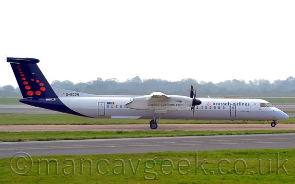 Side view of a white, high-winged, twin propellor-engined airliner taxiing from left to right.
The plane has a grey lower half, with a dark blue, almost purple, rear fuselage and tail, with a series of red dots in the form of a lower-case letter "b" on the tail.
There are black "Brussels Airlines" titles on the upper forward fuselage, the web address "brusselsairlines.com" on the engine pods slung under the wings, and the registration "G-ECOH" on the upper rear fuselage.
A strip of green grass stretches across the foreground, with more grass defining the edges of a couple of taxiways and a runway in the background.
Trees in the distance line the horizon, under bright but hazy skies.