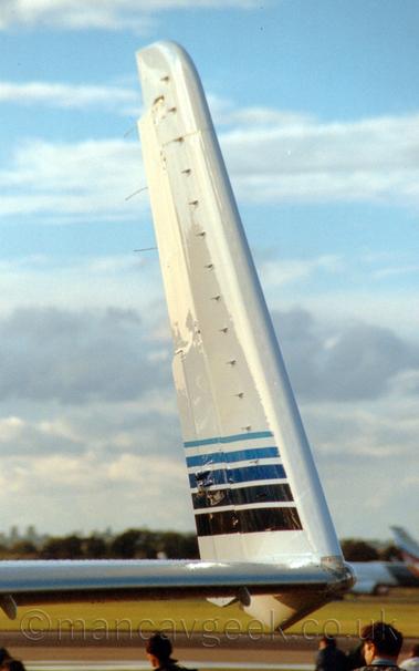 Close-up of the inside face of the left upturned wingtip of a white BizProp parked facing to the right.
There are 5 blue stripes running from front to back, pale blue and thin at the top, getting darker and thinner as they get lower, the lowest one being almost black.
The heads of people walking around in the background can be seen at the bottom of the frame.
A couple of out-of-focus planes can be seen in the distance, with trees behind them stretching across the lower part of the frame, the rest being filled with streaks of pale blue sky and clouds.