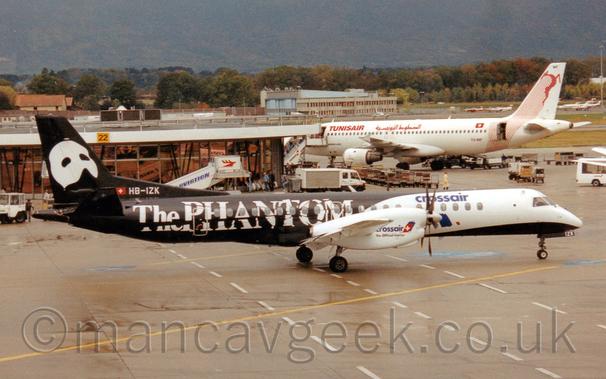 Side view of a white and black, twin propellor-0engined airliner taxiing from left to right through a busy airport scene.
The plane has a white forward fuselage with a black belly, and white engine pods on the wings with blue "Crossair" titles in front of the wings.
The rear fuselage is black, with large white "The Phantom" titles in a fractured font.
The black tail has a large, white mask that would cover the top half of someones face, the lower part of the nose and the mouth being uncovered.
The registration "HB-IZK" is on the lower part of the tail in white.
Grey apron fills the foreground, with a larger, white and red twin engined jet airliner on the right of the frame parked next to a circular terminal building on the right.
White trucks, vans, and mobile airstairs litter the background, along with rows of open baggage carts.
A variety of light aircraft and bizjets are parked on the far side of the runway in the distance on the right of the frame, with a large grey concrete hangar in the middle, with trees stretching across the frame beyond that, with a tree covered mountain rising to fill the rest of the frame at the back.