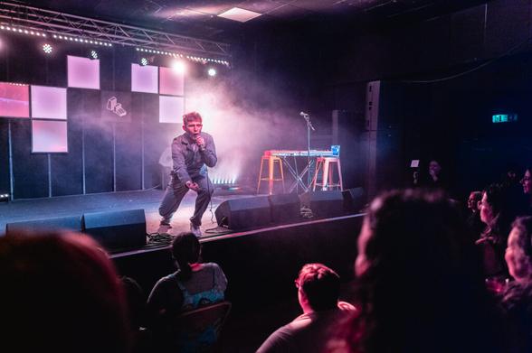 A performer on stage sings into a microphone under colorful lighting, surrounded by smoke, with a keyboard and stools nearby. Audience members watch from their seats in the foreground.