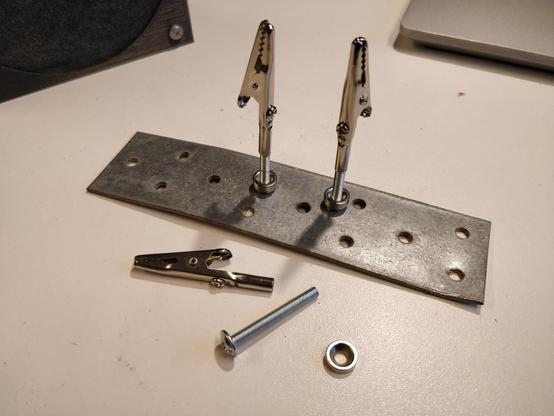 A photo of an improvised set of "helping hands" for soldering.

Two alligator clips, each attached to a screw, stand on top of a small nail plate with the help of small magnets attached to the screws.

On the table, in front of the nail plate, is an alligator clip, a screw and a small magnet with a hole through it. This is to show of the parts that make up the helping hands.
