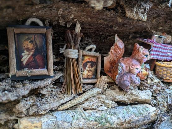 Image of a fallen, gnarled log, with a crevice that someone has adorned with a pair of small squirrel figurines and decorations. This photo is a closeup of two miniature paintings, both exquisitely detailed and in a realistic style. One is of a regal-looking squirrel wearing a noble's red petticoat and an ascot, the other of a squirrel wearing a white hoop skirt dress and holding an orange parasol.