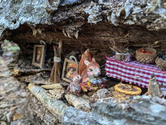 Image of a fallen, gnarled log, with a crevice that someone has adorned with a pair of small squirrel figurines and decorations. This photo is frontal view of the squirrel figurines. One appears to be a parental figure  with rosy cheeks holding a vibrant red, yellow and green leaf as a food platter, with several small, sculpted baked goods on top. The other squirrel figure is right next to them, much smaller and holding their own baked good and smiling joyously.