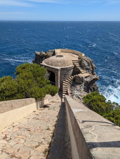 A stone walkway lined with trees leads down to a domed structure on a rocky outcrop overlooking a rough but very blue sea.