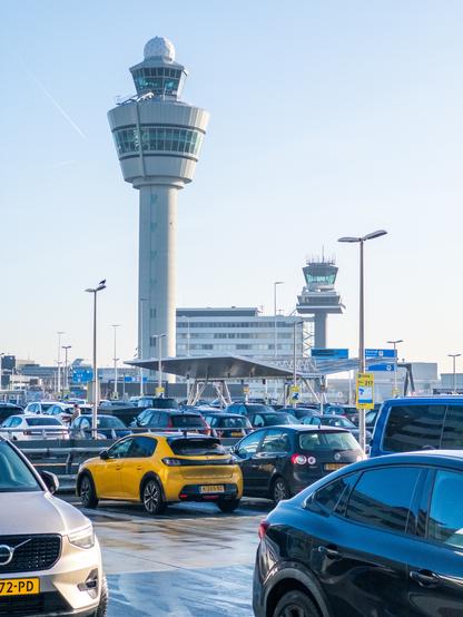A busy parking area with parked cars and an airport control tower at Schiphol, Netherlands.