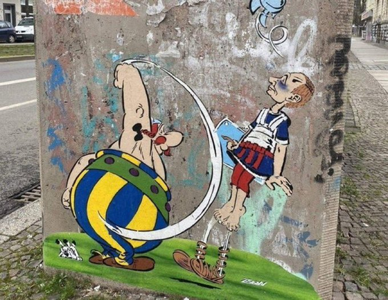 Auf einer Betonwand hat ein anonymer Künstler ein Graffiti gemalt. Obelix, dessen blau-weiße Hose nun die ukrainischen Farben hat, schlägt einen Legionär, der Putin darstellen soll.