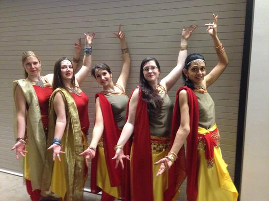 5 women posed with one arm up , one arm down in Bollywood dance pose. 
Two wear red tops with gold dupattas, three wear gold tops with red duppatas. All wear yellow  satin over skirts and red coin scarves.