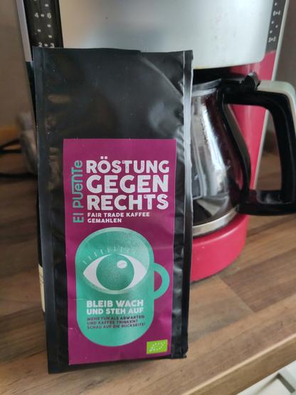 Vor einer Filterkaffeemaschine mit Glaskanne steht eine schwarze 250g-Packung Kaffeepulver. Auf dem lila Etikett ist ein grüner Kaffeebecher abgedruckt. Darüber steht groß "Röstung gegen Rechts", daneben der Markenname "El Puente" und darunter steht "Faire Trade Kaffee gemahlen". Auf der Tasse steht "Bleib wach und steh auf" und "Mehr tun als abwarten und Kaffee trinken? Schau auf die Rückseite."