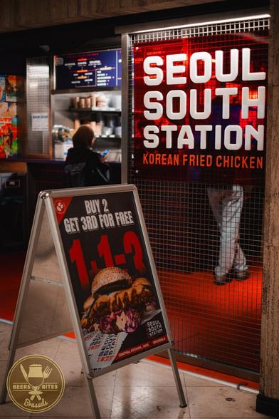 A restaurant named "Seoul South Station" advertises Korean fried chicken and a "buy 2, get 3rd free" deal. An A-frame board outside shows an image of a Korean burger. A logo for “Beers and Bites Brussels” is visible.
