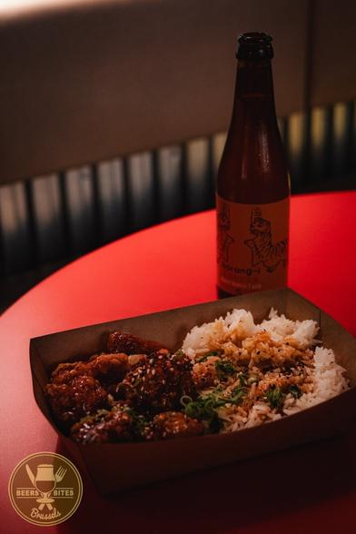 Cardboard tray with fried chicken, white rice, and garnish, on a red table. A bottle of beer sits behind it labeled “Horangi.” A logo for “Beers and Bites Brussels” is visible.