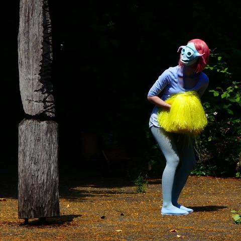 Ein Bild einer Person mit rosa Haaren, die eine hellblaue Maske trägt, die einem außerirdischen Wesen ähnelt. Sie trägt ein hellblaues Oberteil und hellblaue Strumpfhosen mit einem gelben Pompon in der Hand. Sie steht vor einem alten, grauen Holzpfosten in einem Garten mit etwas Grün im Hintergrund.