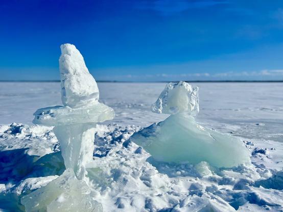 Two ice sculptures stand on a snowy landscape under a clear blue sky. The sculpture on the left resembles a tall, pointed structure, while the one on the right has a more rounded shape, possibly resembling a figure. The surrounding area is covered in snow, creating a serene winter scene.