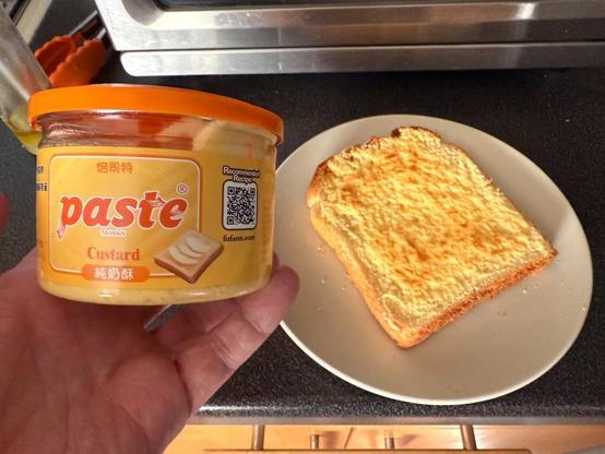 A hand holding a plastic jar. The jar has an orange lid and an orange theme to the label. It is simply cold “paste“ underneath that it indicates that it is from Taiwan and that it is custard. There are Chinese characters on here.

In the background is a small plate with a piece of toast covered in paste.