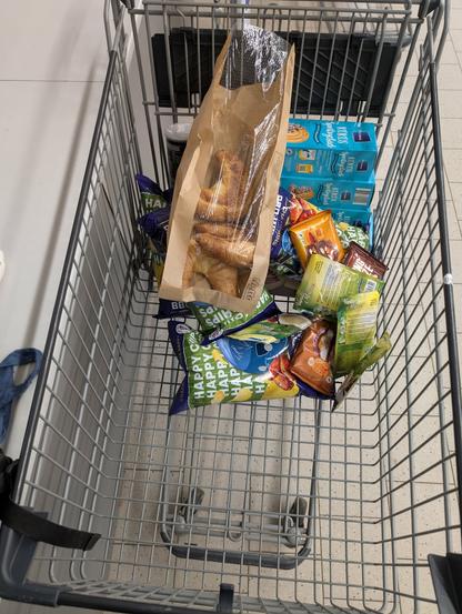 Alt-text von ChatGPT aus Basis des Fotos und manueller Angaben:

Ein silberner Einkaufswagen mit schwarzen Kunststoffelementen steht in einem Supermarktgang. Darin liegen mehrere Lebensmittel, vor allem auf einer Seite konzentriert. Auffällig ist eine große braune Papiertüte mit durchsichtiger Plastikfolie, gefüllt mit goldbraunen Croissants.

Darunter und daneben befinden sich zahlreiche Snacks. Mehrere Tüten „Happy Chips“ in grüner und blauer Verpackung sind sichtbar, mit Sorten wie gesalzen, BBQ und Paprika. Die Tüten liegen ungeordnet im Wagen. Außerdem sind einige Süßigkeiten dabei, darunter mehrere blaue Packungen mit der Aufschrift „Keks“, die knusprige Kekse mit einer Füllung zeigen.

Zusätzlich gibt es weitere kleine, bunte Snackpackungen und ein schwarzer Becher ragt am oberen Rand des Wagens heraus. Der Einkauf besteht überwiegend aus süßen und salzigen Snacks, möglicherweise als Trost oder Belohnung.

Der Einkaufswagen ist für die Menge an Produkten zu groß, was darauf hindeutet, dass der Einkauf spontan war. Der helle Supermarktboden mit großen, quadratischen Fliesen ist im Hintergrund sichtbar.