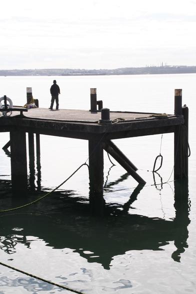 Beeld van een man met hoed op het uiteinde van een steiger, die een hoek maakt, donker silhouet. 
De steiger wordt weerspiegeld in het water. In de verte een vage landtong van Ierland.