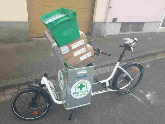 Vélo cargo douze cycles de la pharmacie du vélo, de Strasbourg.
Roue 406 devant 559 derrière.
Je l'appelle la Ferrari de la pharmacie. Bon Linhof serait mieux, marre de citer des métallosaures.
Vélo léger, rapide, maniable, qualitatif, avec une belle caisse en alu devant, sur laquelle est écrit: "Pharmacie du vélo".
Le logo est un jeu de mots visuel, sorte de rébus, avec un plateau de vélo et ses dents entourant la croix verte des pharmacies.
Bon revenons au sujet: la caisse du vélo déborde avec deux fois son volume. Seul son couvercle maintient précairement cartons et cageots.
À un point où c'est un peu dur de voir la route...!
Faut rouler doucement quoi.
Derrière, on voit trottoir et maisons et le vélo est à l'arrêt avec sa béquille double, une paire de gants est posée sur la selle.